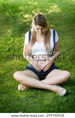 Pregnant young woman relaxing on grass and holding hands at shape of heart on abdomen