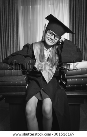 Black and white portrait of smart girl in graduation cap posing on table at library