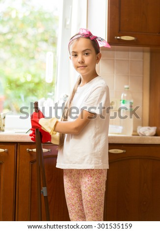 Portrait of smiling girl posing on messy kitchen with broom and scoop
