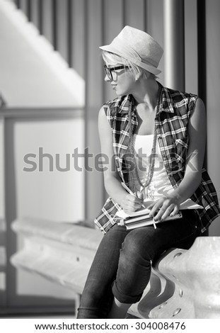 Black and white portrait of stylish girl in eyeglasses and hat writing in notebook
