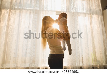 Cheerful mother playing with her baby son against big window in sun rays