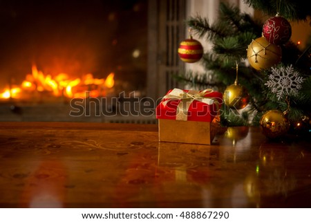 Closeup image of red gift box on wooden table in front of burning fireplace and Christmas tree