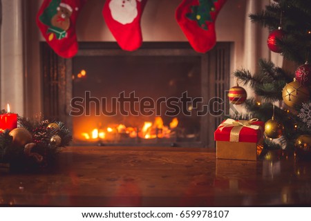 Closeup of red Christmas gift box with golden ribbon next to decorated Christmas tree and fireplace