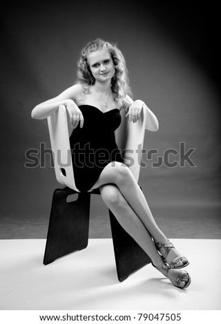 Black and white photo of young curly blond lady sitting on white chair in evening dress