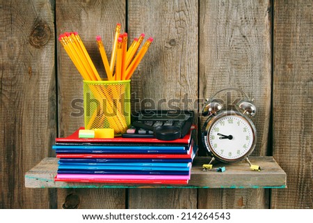 Writing-books, an alarm clock and school tools on a wooden shelf. On a wooden background.