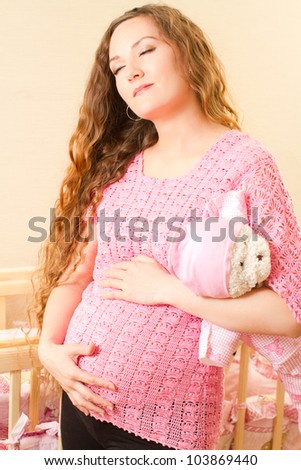 Pregnant woman with long hair and toy Teddy bear near a crib