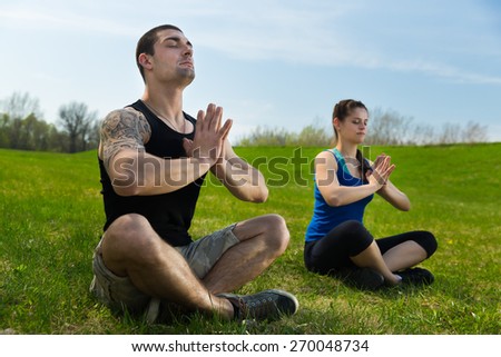 Young man together with young woman is doing yoga exercises and sitting on grass .