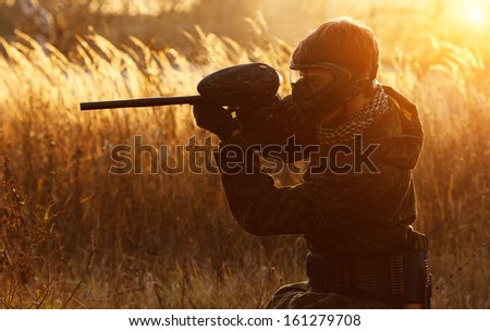 Paintball sport player in protective uniform and mask aiming gun before shooting at sunset