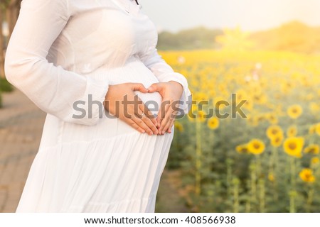 Pregnant woman with heart sign on her belly and sun flower garden background.