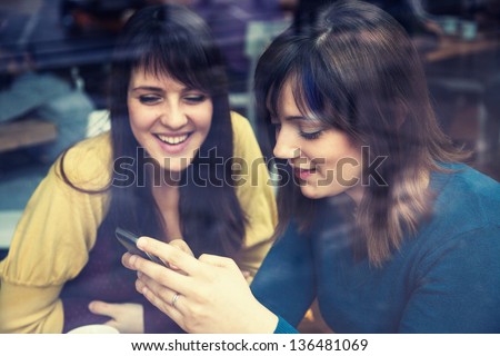 Two Young Girls Smiling Using Smart Phone In A Cafe. The Photo Was Taken Through The Window
