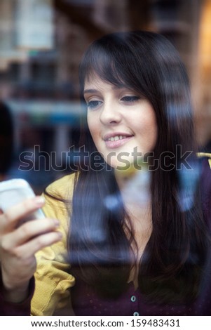 Beautiful smiling girl using smart phone in a cafe. The photo was taken through the window