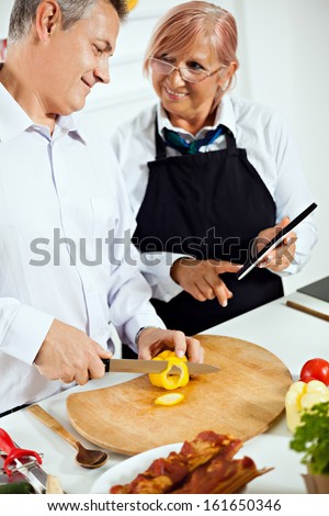 Happy mature couple cooking together in kitchen. They are reading the recipe using tablet.