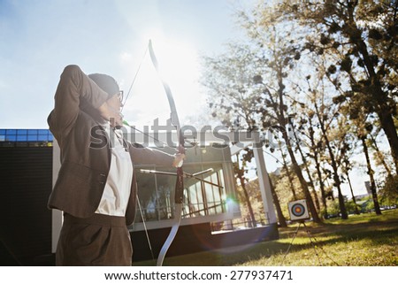 Businesswoman Pointing The Target With Bow And Arrow In Front Of Business Building
