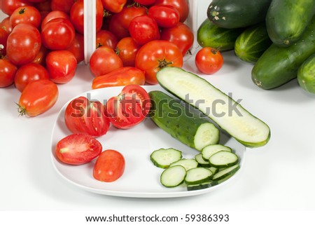 A detail studio view of two sliced field tomatoes and a cucumber and a basket of ripe field tomatoes and a bunch of cucumbers in the background.