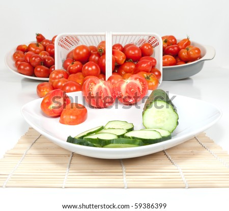 A wide angle studio view of two sliced field tomatoes and a cucumber and a basket and dishes of ripe field tomatoes with the main focus on the two sliced tomatoes on the plate.