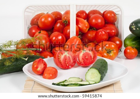 An front detail studio view of plated sliced tomatoes and cucumber with a basket of ripe field tomatoes and a sprig of dill and a few cucumbers in the background.