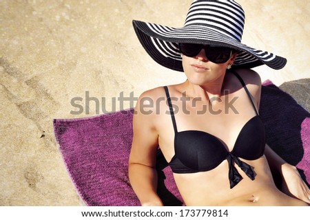 Young glamorous woman in a summer hat, sunglasses and a black bikini laying on the beach.