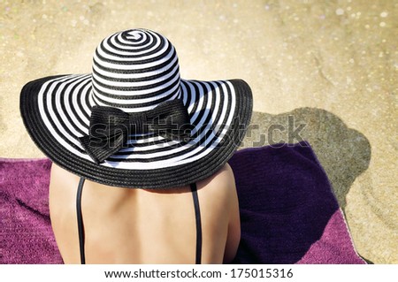 Young elegant woman wearing a black bikini top and a black and white striped beach hat sunbathing on the beach.