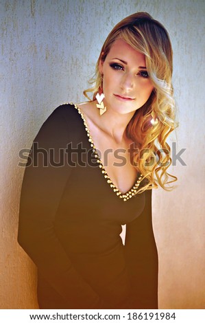 Beautiful young woman with curly blonde hair and smoky eye makeup wearing a black long sleeved dress with golden studs and matching big golden earrings.