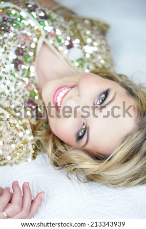 Beautiful young happy woman with shoulder length curly hair wearing a golden sparkly sequin bolero and pink evening makeup is lying down on the bed and smiling.