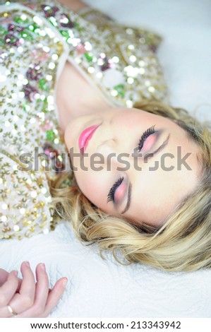 Beautiful young blonde woman with shoulder length curly hair wearing a golden sparkly sequin bolero and pink evening makeup is lying on the bed with her eyes closed.