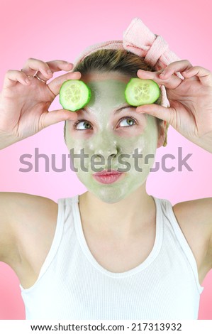 Beauty portrait of a happy young woman wearing a pink headband , white tank top and a green facial mask is holding two slices of cucumber above her eyes. Pink background.