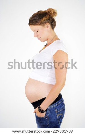 Young pregnant woman with dark hair wearing jeans and a white crop top is standing in profile and looking down at her growing baby bump.