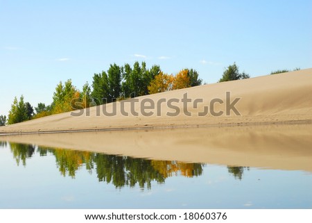 A glassy lake mirrors a sand dune with trees turning to their fall colors behind it.