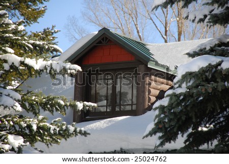A snow covered cabin\'s dormer peeks out between two pine trees.