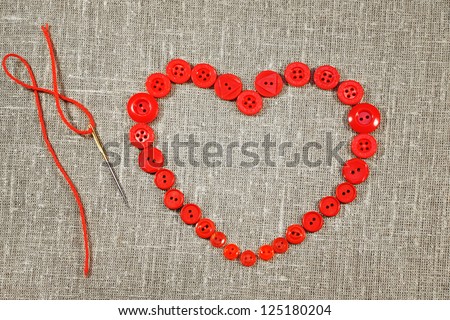 Heart in shape of red buttons and darning needle. Objects on unbleached fabric.
