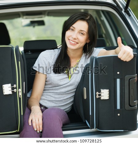 Young female sitting in the trunk of a car with suitcases, showing thumb up sign, ready to leave for vacations