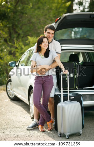Young couple standing near the opened car boot with suitcases, looking to the camera, outdoors