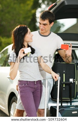 Young couple standing near the opened car boot with suitcases, holding blank credit cards, looking at each other, outdoors
