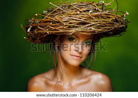 Closeup of beautiful young woman with fresh makeup and wicker nest on her head over green background