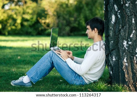 Young man sitting near the tree stem and using laptop outdoors, looking to the side