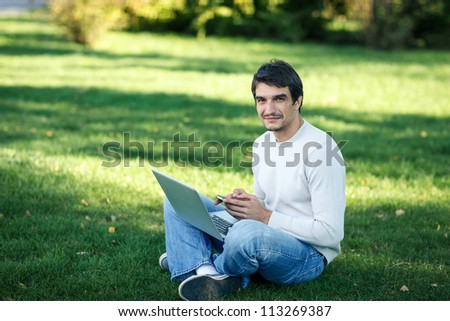 Smiling young man with laptop outdoor sitting on the grass and chatting on the cellphone