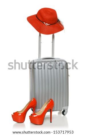 Silver suitcase with red straw hat on the handle and red high heel shoes near it, isolated on white background