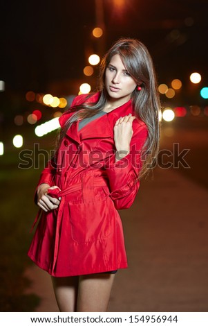 Fashion woman in red trench coat posing against evening street lights background
