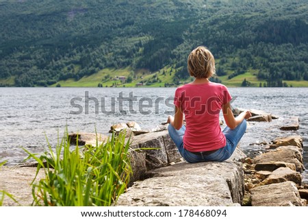 Woman relaxing at lake shore enjoying summer freedom meditating in lotus position, Norway. Caucasian girl on summer travel holidays vacation outside.