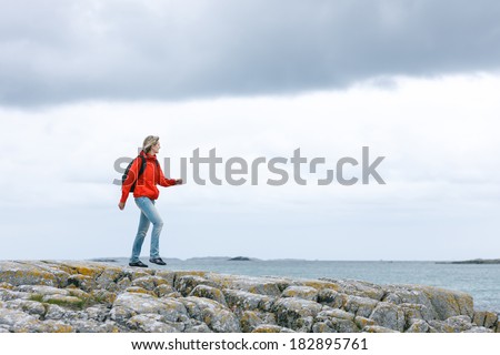 Woman hiker in red jacket and backpack walking on rock cliff against sea / ocean background