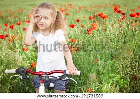 Happy smiling little girl standing with bicycle on the poppy meadow looking away through circled fingers