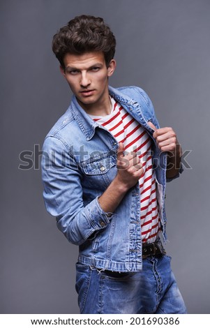Man fashion model, stylish young man standing posing in denim holding his jacket, over gray background