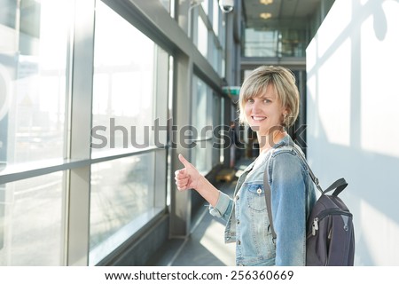 Happy woman with backpack standing in airport building going on boarding, gesturing thumb up