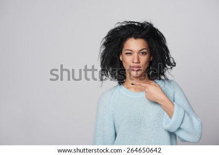 Funny winking multiracial african american - caucasian girl pointing at blank copy space next to her, over gray background