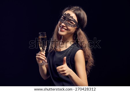 Happy sensual woman wearing party mask, holding a glass with champagne over dark background and showing thumb up gesture.