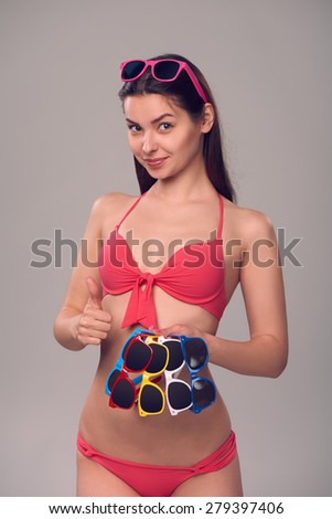 Smiling woman in pink bikini holding many colourful sunglasses, over gray background