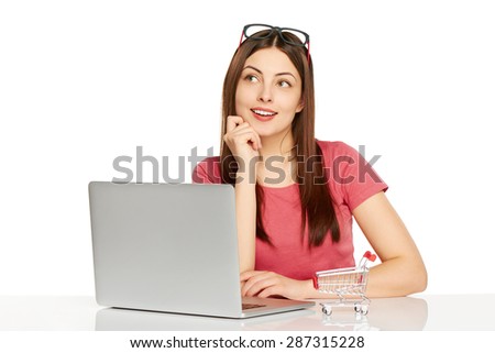 Online shopping concept. Smiling thinking woman sitting at table with laptop, small empty shopping cart standing on table, isolated on white background