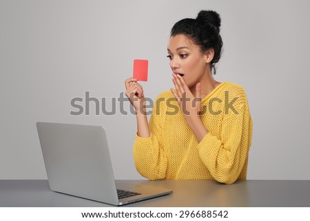 Surprised mixed race african american - caucasian woman with laptop computer holding blank credit card, sitting at table, over gray background