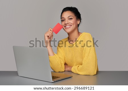 Happy laughing mixed race african american - caucasian woman with laptop computer showing blank credit card, sitting at table, over gray background