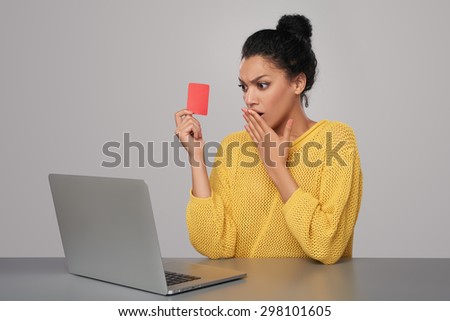 Shocked mixed race african american - caucasian woman with laptop computer holding blank credit card, sitting at table, over gray background